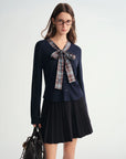 Woman wearing a navy blue long-sleeve top with a plaid bow and black pleated skirt on a white background