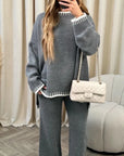 Person wearing a gray sweater and pants with a white quilted handbag, standing in a room with curtains and a chair.