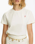 Person wearing a white t-shirt with a small red logo and gold belt with charms on a plain background