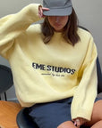 Person wearing a yellow 'EME STUDIOS' sweater sitting on a chair.