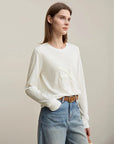 Woman wearing a white sweater and blue jeans with a beige background, featuring the brand 'Amii'.