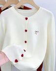 White sweater with red heart buttons held by a hand against a neutral background