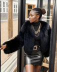 Woman wearing a black fur coat and leather skirt standing by a glass door.